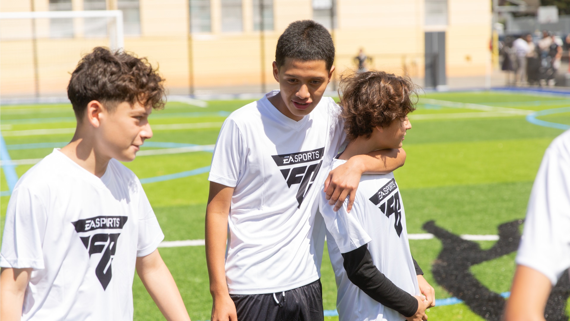 Three youths in white sports shirts stand together on a sunny artificial turf soccer field, with one resting an arm on a teammate's shoulder.