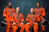 Four astronauts in orange spacesuits pose holding black helmets against a dark background.
