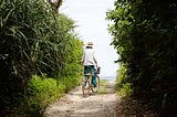 A person in a straw hat, white blouse, and blue tie-dye pants rides a bicycle with a front basket along a narrow, sun-dappled dirt path lined with lush green tropical foliage, heading toward a glimpse of the ocean in the distance.