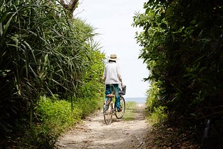 A person in a straw hat, white blouse, and blue tie-dye pants rides a bicycle with a front basket along a narrow, sun-dappled dirt path lined with lush green tropical foliage, heading toward a glimpse of the ocean in the distance.