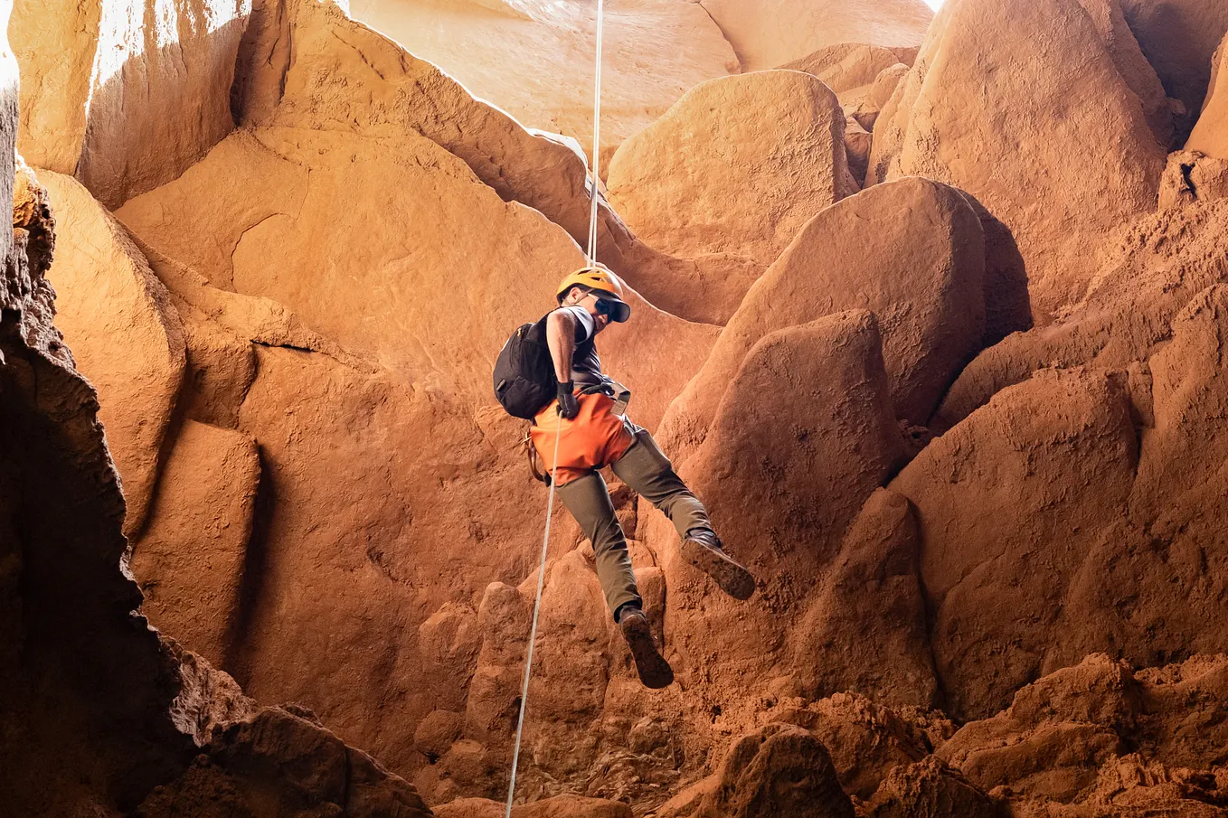 A person wearing an orange harness, yellow helmet, and backpack rappels down a rope through a narrow sandstone canyon with smooth, curved orange and red rock walls.”