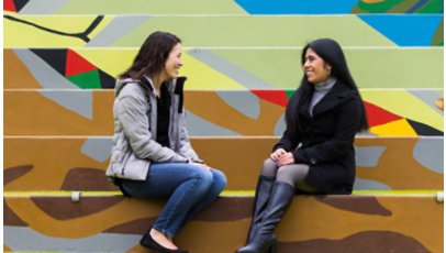Two women sit outside on stairs while talking.