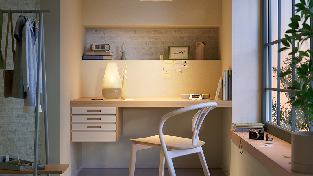 An office with a desk, chair, lamp and office supplies, with a window, hanging clothes and a potted plant on either side