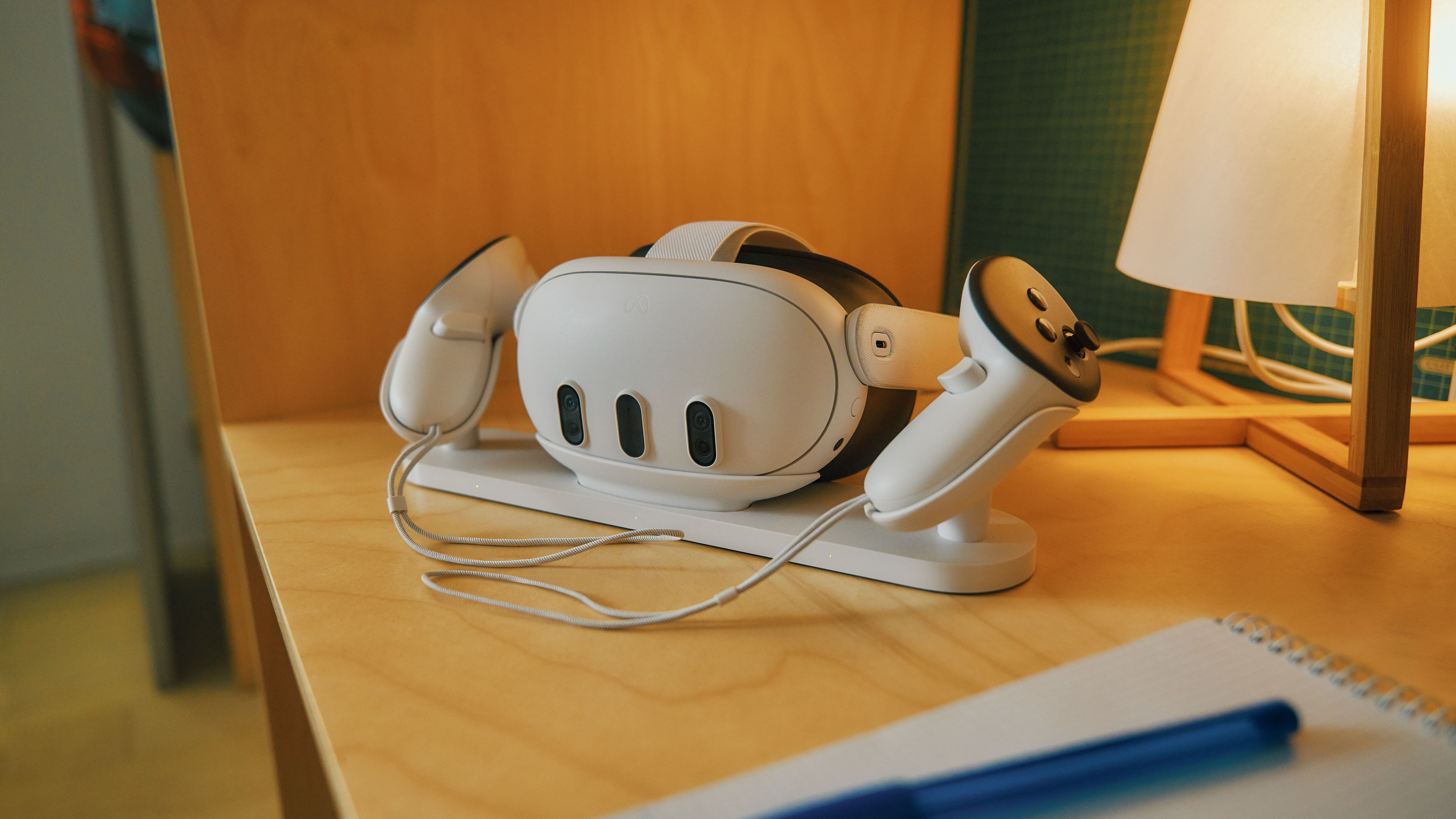 The Meta Quest 3 and controllers on their charging station, which is itself on a wooden desk next to a lamp