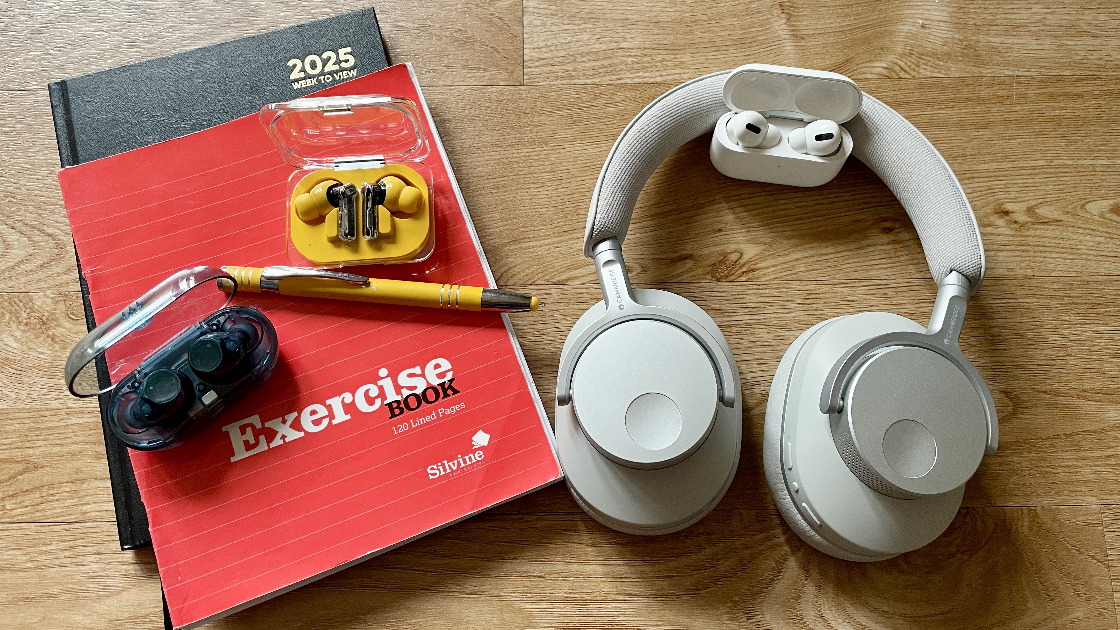 The Cambridge Audio Melomania P100, Sony WF-C710N, AirPods Pro 2 and Nothing Ear (a), which all feature in this student headphones guide, on a wooden table next to a stack of exercise books, a diary and a pen