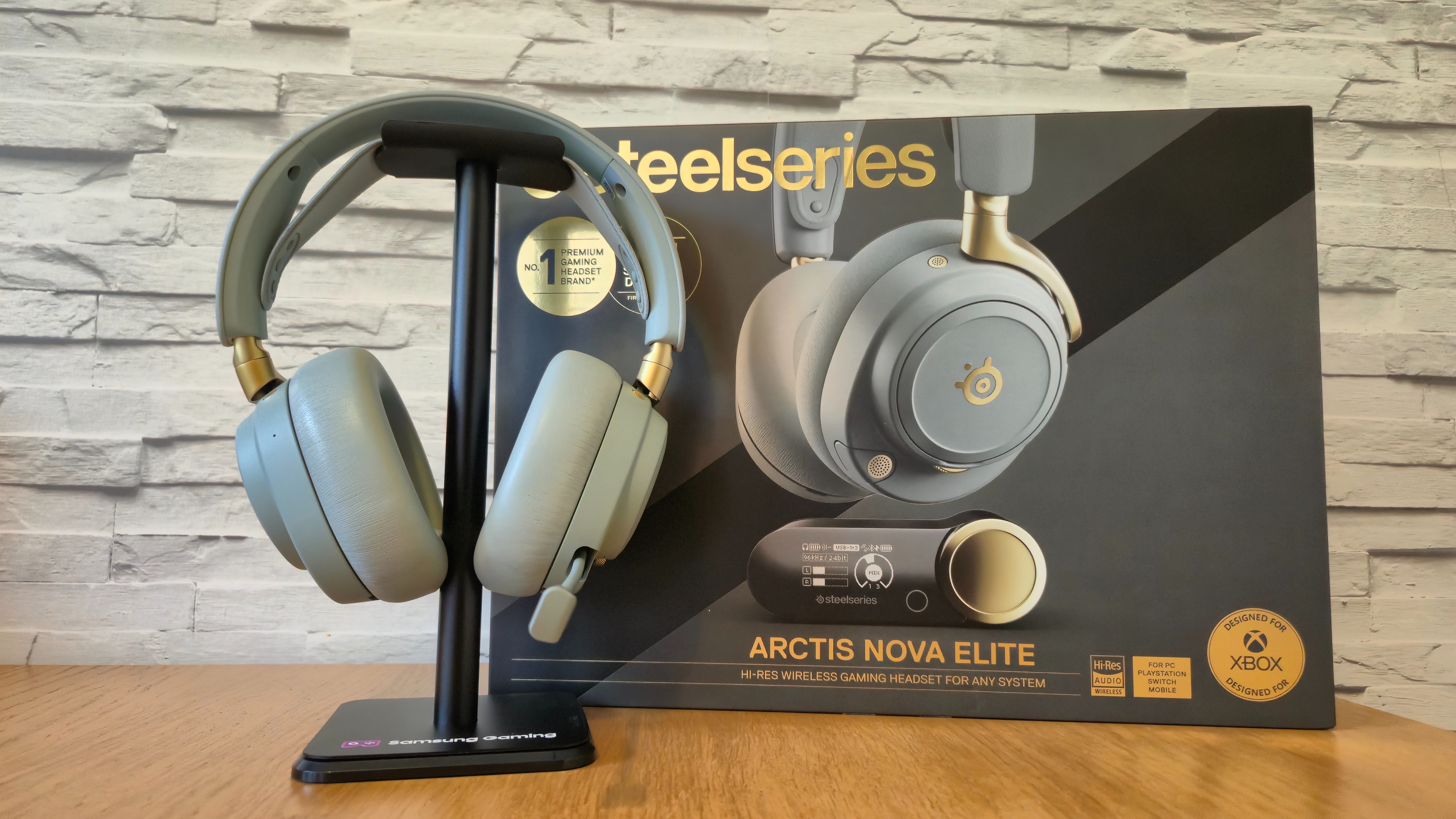 The SteelSeries Arctis Nova Elite headset on headset stand in front of a white brick background and on a wooden surface