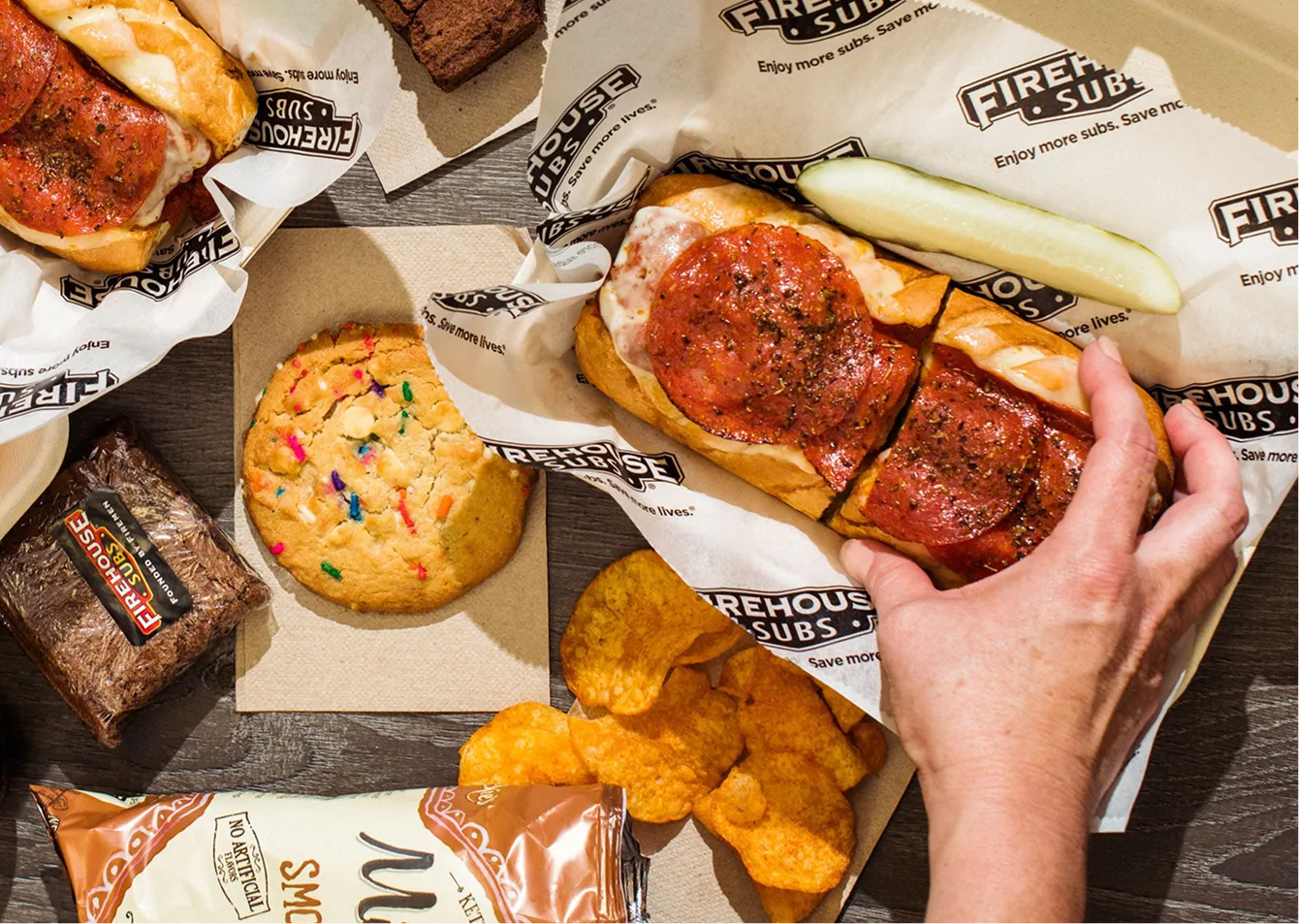 Hand grabbing a pepperoni sub sandwich wrapped in Firehouse Subs paper with a pickle, chips, cookie, and brownie on a wooden surface.