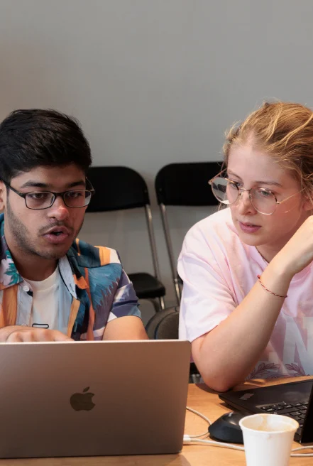 Two people sitting side by side at a table, collaborating while looking at a laptop screen.