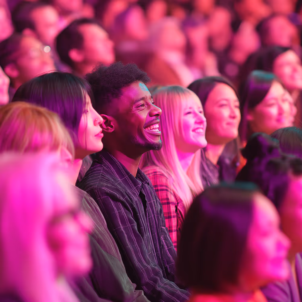 Diverse audience attentively watching and smiling during an event under pink stage lighting.