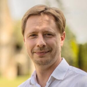 Emmanuel faces the camera with a slight smile. The background is blurred, but it actually is a gorgeous abbey.