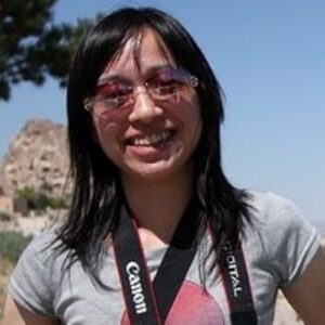 A headshot of Patricia smiling, carrying a camera, and wearing a grey t-shirt and sunglass.