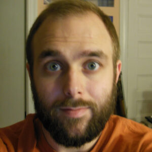 The photograph is an old selfie of John wearing an orange shirt. The background contains two doors and his head blocks a poster of a space shuttle launch.