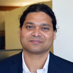 Sameer Patil looking at the camera and smiling against a faded background of walls in an office space.