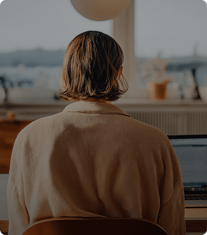 A person with short hair is sitting at a desk in a softly lit room, working on a laptop.