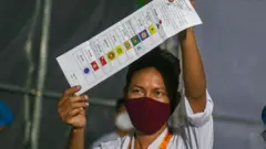 Một nhân viên bầu cử đeo khẩu trang đỏ giơ phiếu bầu trong quá trình kiểm phiếu vào ngày bầu cử Quốc hội Myanmar năm 2020