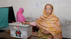 একজন নারী ভোটের বাক্সে ব্যালট ফেলছেন, পেছনে দাঁড়িয়ে আছেন আরো একজন নারী