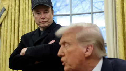 Musk hovers in the background behind Donald Trump in the White House Oval Office