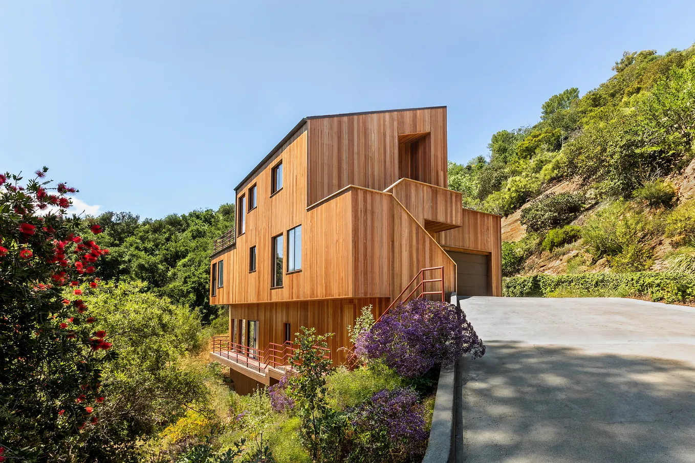 A contemporary, multi-level home nestled on a steep, vegetated hillside. The exterior is completely clad in light brown vertical wood siding. A curved concrete driveway leads up to a garage on the right side of the house. The home features dark-framed windows, a lower-level balcony with red railings, and is surrounded by vibrant green trees and purple and red flowering bushes on a sunny day.