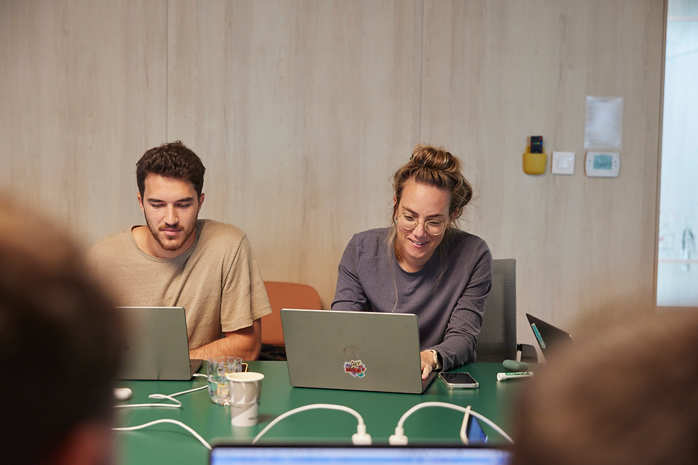 Wix employees in a meeting room working on their laptops. A man in a beige t-shirt and a woman in glasses and a dark sweater are seated at a green table, both focused on their screens.