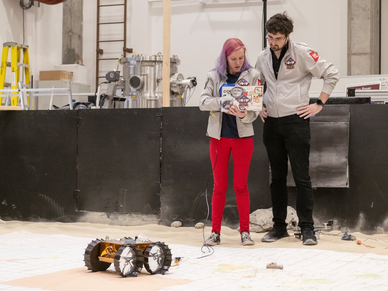 Two people consult a laptop in a room made to resemble the moon's surface while a lunar lander waits in the foreground.