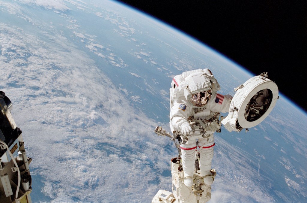 An astronaut in a white spacesuit works on a fixture while outside of the International Space Station. He holds a large white circular object in his left hand. Earth can be seen behind him: the planet's blue water and white clouds take up most of the background, but a sliver of black space can be seen at top right.