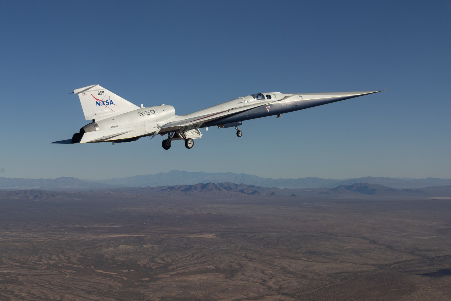 The X-59 quiet supersonic research aircraft flies above California. The plane has a distinctive shape with a long, sharply pointed nose. The nose is silver, while the rest of the body looks white. The words "NASA" and "X-59" are on the body of the aircraft. In the background, we can see brown earth below the plane as well as hazy mountains in the distance.